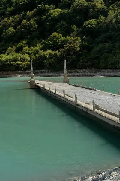 A narrow concrete pier with stone pillars extends into turquoise lake water, with forest on the opposite shore.