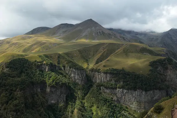 A panoramic view of a mountain valley with steep rocky slopes, green meadows, and cloudy sky above.