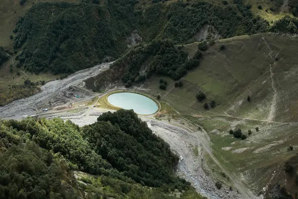 A small turquoise mountain lake lies in a green valley, surrounded by steep hills, trees, and rocky slopes.