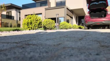 Lüks bir villanın önünde kalkışa hazırlanan bir İHA 'nın 4K hava görüntüsü. Kapısı açık bir araba, yeşil çimenler ve yürüyen insanların gölgeleri. Lüks, teknoloji ve emlak projeleri için mükemmel..