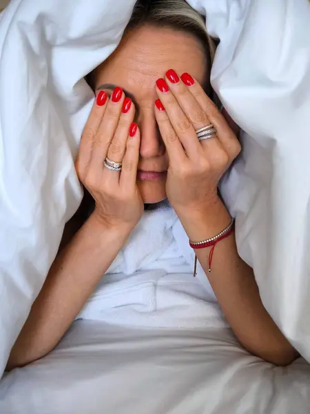 Parlak manikürlü bir kadın yüzünü ellerine saklar, beyaz bir battaniyenin altından dışarı bakar. Yüz ifadesi gizlidir, ama poz güçlü duygular taşır..