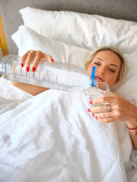 Genç çekici sarışın kadın beyaz yatakta uzanıyor şeffaf şişeden cam bardağa temiz su döküyor. Sabah ritüeli, sağlıklı yaşam tarzı, susuzluk.