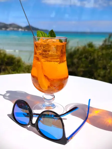 Serinletici yaz kokteylleri beyaz masada servis edilir. Güneş gözlükleri yakınlarda durur. Arka planda okyanus manzarası ve yeşil tepeler bulanıktır.