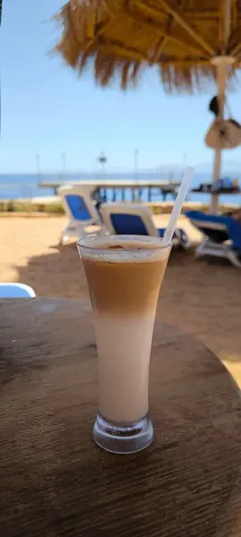 Süt ve buz dolu uzun bir bardak şeffaf bir kamışla ahşap bir masanın üzerinde duruyor. Mavi bir gökyüzü, deniz, güneşlenme yerleri ve kumlu bir plaj.