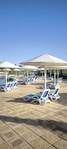 Mavi şilteli iki rahat güneş şezlongu havuzun yanındaki büyük beyaz şemsiyenin altında duruyor. Rahatlamak için ideal bir yer.