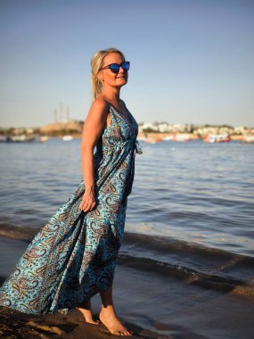 Uzun mavi yaz elbiseli ve güneş gözlüklü zarif kadın gün batımında kumlu sahilde çıplak ayakla ayakta duruyor.