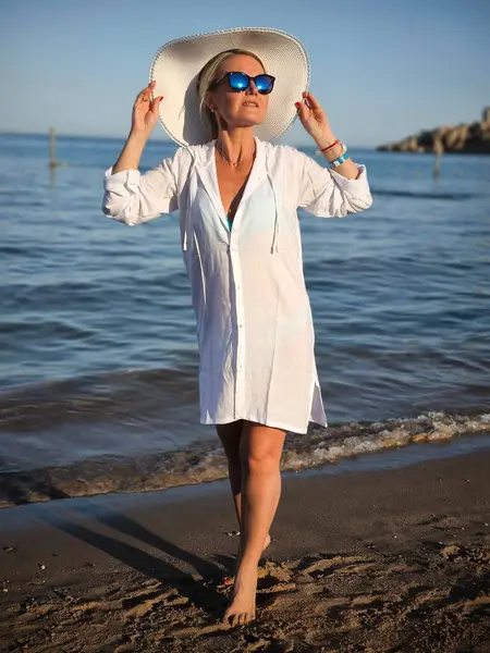 Beyaz plaj ceketli, geniş kenarlı hasır şapkalı ve güneş gözlüklü zarif kadın gün batımında kumlu sahilde yalınayak ayakta duruyor.