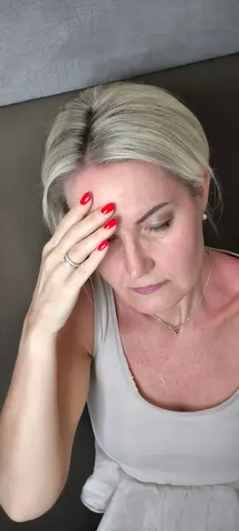 Sarı saçlı yetişkin bir kadın, şakağa el ele tutuşuyor, parmaklarını alnına bastırıyor, acı, yorgunluk ya da stres ifade ediyor, yüzü endişeli ve düşünceli.