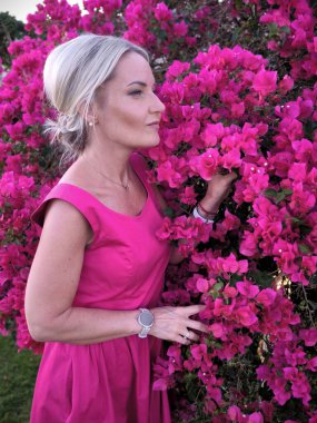 Sarı saçlı, parlak pembe elbiseli bir kadın geniş bir fuşya begonvili çalılığın yanında duruyor.