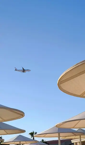 Güneşli bir günde, bir yolcu uçağı mavi gökyüzünde, plaj şemsiyeleri ve dinlendirici bir atmosfer yaratan güneş şezlonglarının üzerine iniyor..