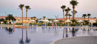 Küçük mavi mozaik fayanslarla kaplı açık havada yüzme havuzu güzel bir desen oluşturuyor. Havuz, açık gökyüzünü ve kıyı boyunca uzanan uzun palmiye ağaçlarını yansıtır.