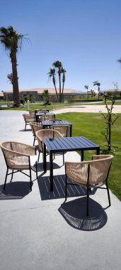 Açık teras, kafe veya restoran hasır bahçe mobilyaları, mobilyalar yüksek palmiye ağaçları, çimenler ve açık mavi gökyüzüne karşı beton kaldırım üzerinde yer almaktadır.