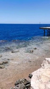 Küçük bitkiler ve kumlu bir patika ile kayalık bir kıyıya yakın denizin masmavi sularına uzanan ahşap bir iskele manzarası.