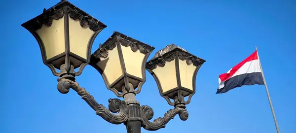 Rüzgarda dalgalanan Mısır bayrağının yanında açık mavi bulutsuz bir gökyüzüne kurulmuş üç lambadan oluşan klasik bir lamba.