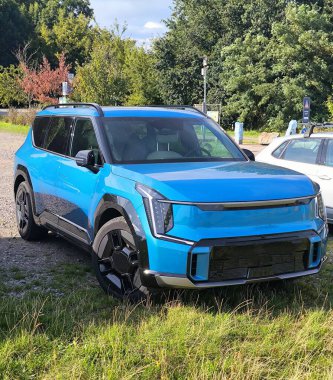 Parlak mavi bir Kia EV9 elektrik geçidi yeşil çimlerin yanındaki toprak yolun kenarına park edilmiş.