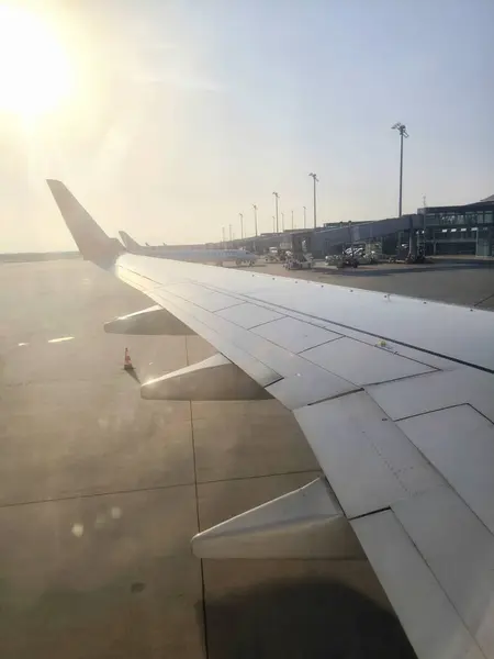 Güneşli bir günde kanadın uçak penceresinden ve havaalanı önlüğünden bakın. Parlak güneş, pistin betonundan yansıyarak kanadın yüzeyini aydınlatır ve parlar.