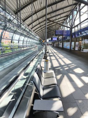 Leipzig, Almanya - 13 Ağustos 2025: Modern bir havaalanında bir sıra boş sandalye. Koltukların yanında metal bir çöp kutusu duruyor. Hareket eden yürüyen merdivenler solda görünüyor.