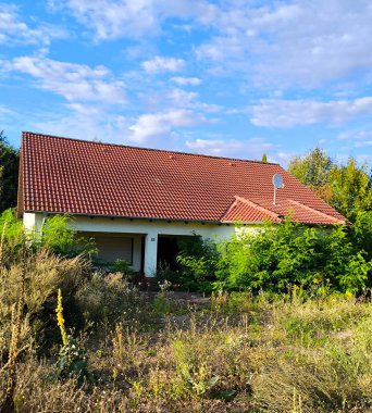 Terk edilmiş bir ev, uzun otların ve çalıların arasında gömülü. Kırmızı kiremitli çatı beyaz duvarlarla çelişiyor, terk edilme ve melankolik hissi yaratıyor..