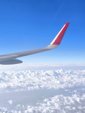 Sonsuz gökyüzünün ve kabarık bulutların uçak penceresinden bak. Ön planda uçağın kanadı ve ucunda parlak kırmızı bir element var.