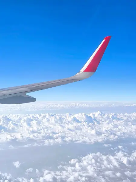 Sonsuz gökyüzünün ve kabarık bulutların uçak penceresinden bak. Ön planda uçağın kanadı ve ucunda parlak kırmızı bir element var.