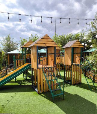 A modern playground on a sunny summer day. The play complex features two wooden play structures and various slides, ladders, and fenced platforms, set on an artificial green surface.