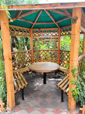 A cozy wooden gazebo with a hexagonal shape, located in a shady garden. Constructed of light wood, the gazebo features a round wooden table and built-in benches for relaxation.
