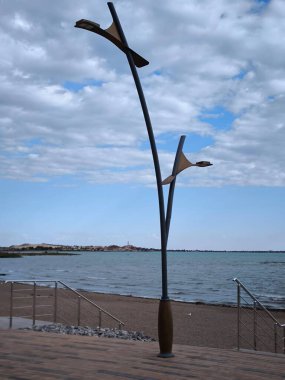 A photo of a modern promenade with a multi-colored tiled sidewalk leading to the beach and a tall, minimalist designer street lamp resembling a tree or branch, against a backdrop of blue sky and sandy beach