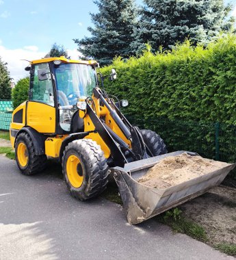 The construction loader is positioned on a paved road, its bucket lowered and filled with sand. Heavy-duty, deep-tread tires and a brightly coloured cabin highlight its intended use in construction, roadwork, and landscaping.