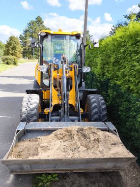 The construction loader is positioned on a paved road, its bucket lowered and filled with sand. Heavy-duty, deep-tread tires and a brightly coloured cabin highlight its intended use in construction, roadwork, and landscaping.