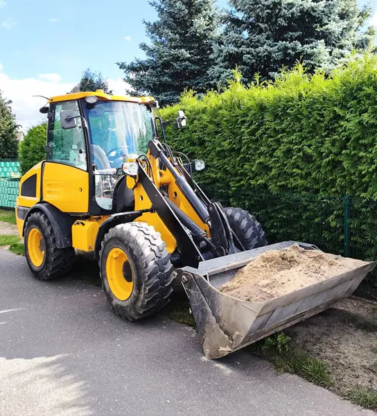 The construction loader is positioned on a paved road, its bucket lowered and filled with sand. Heavy-duty, deep-tread tires and a brightly coloured cabin highlight its intended use in construction, roadwork, and landscaping.