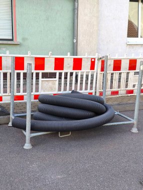 A coil of black corrugated pipe for cable protection or drainage systems sits on a metal frame resting on asphalt. The coil is enclosed by red-and-white striped construction barriers.