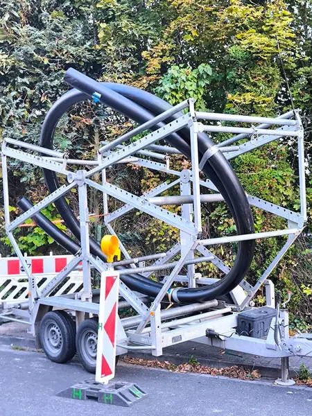 A trailer parked on the side of the road, with the remaining frame holding a large coil of black polyethylene pipes for plumbing, gas lines, or cable lines. The pipes are large in diameter and coiled into a ring.