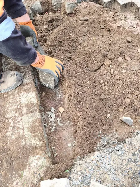 A workers hands, wearing orange protective gloves, are laying curbstones or paving slabs. They carefully place a rectangular stone into a prepared trench filled with loose soil.