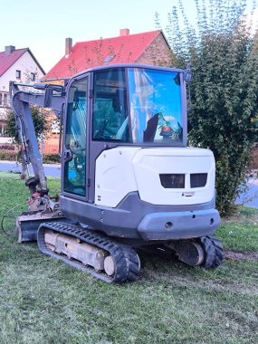 A small, dark gray crawler excavator parked on a lawn in a residential area. The excavator is in working position, with the bucket lowered.