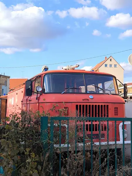 The front of an old vintage fire engine, a bright red IFA W50 model, typical of GDR vehicles. The truck is parked in a parking lot behind a fence covered with dead branches, indicating both its abandonment and its historicity.