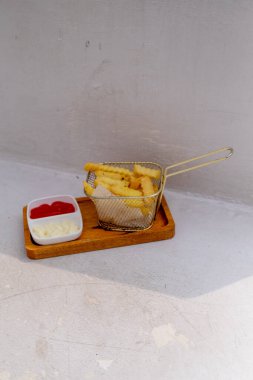 A flatlay photo of French fries served with tomato sauce and mayonnaise, placed in an aluminum basket on a wooden plate. clipart