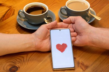 Görüntü iki fincan kahveyle bir masada oturan bir çifti gösteriyor. Elleri birbirine kenetlenmiş ve aralarında ekranda kırmızı kalpli bir akıllı telefon var..