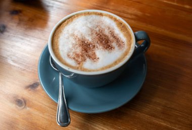 İçinde kaşık olan bir fincan kahve ahşap bir masada duruyor. Kahve krem şanti ile kaplıdır ve üzerinde bir tutam tarçın vardır. Rahatlık ve rahatlama kavramı