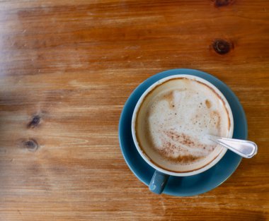 İçinde kaşık olan mavi bir kahve fincanı ahşap bir masanın üzerinde duruyor. Kahve sıcak ve köpüklü, üzerinde birkaç kahverengi benek yüzüyor. Kupa da mavi olan bir çay tabağının üzerine konur.