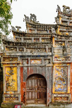 Vietnam 'daki Hue İmparatorluk Şehri' nin yıpranmış, süslü kapısının detaylı görüntüsü. Geçitte karmaşık oymalar, renkli olmasına rağmen canlı oymalar, çiçek ve ejderha motifleri yer alıyor. Tahta kapı biraz aralıktır, içerideki tarihe işaret eder..
