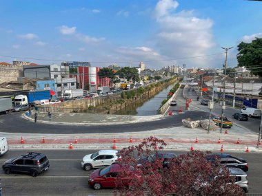 Kenarlarında inşaat konileri olan bir kanala bitişik, arabalar ve kamyonlar da dahil olmak üzere işlek bir caddenin bulunduğu işlek bir şehir manzarası. Gökyüzü parçalı bulutlu. Güneş ışığı ipuçlarıyla dolu. Bu da canlı ve dinamik bir şehir manzarası olduğunu gösteriyor..