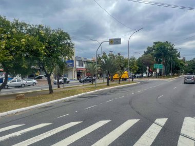 Canlı yeşilliklerle ve bulutlu bir gökyüzü altında sayısız mağaza vitrinleriyle dolu, kentsel yaşam ve ulaşım dinamiklerini resmeden işlek bir şehir caddesi..
