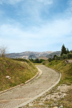 Çimenli tepeler ve dağınık ağaçlarla çevrili asfaltlı, dolambaçlı bir patika, berrak bir gökyüzünün altındaki dingin bir dağ sırasına doğru gidiyor.. 