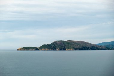 Sakin suları olan sakin bir sahil manzarası, çam ağaçları ve kayalık uçurumlar tarafından çerçevelenmiş, uzak dağlara bakan bir deniz kenarı. 