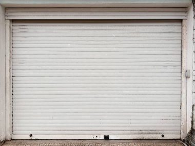 Yatay çizgileri, toprak izleri ve aşınma izleri olan kapalı beyaz metal bir garaj kapısı. Küçük bir renk değişimiyle beyaz bir duvar tarafından çerçevelenmiş.. 