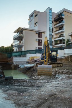 Çağdaş konut binaları ve inşaat malzemeleriyle çevrili, ön planda bir kazıcı bulunan kentsel bir alanda bir inşaat alanı.. 