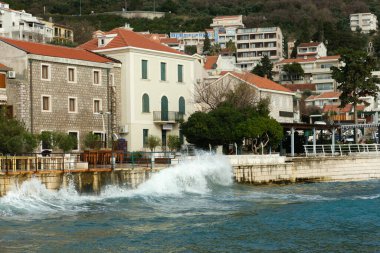 Seafront view of stone and modern buildings with red tile roofs, as waves crash against the shoreline in a coastal town.  clipart