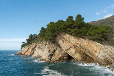 Katmanlı kaya oluşumları ile dramatik bir kayalık ve tepesinde büyüyen çam ağaçları, berrak bir gökyüzünün altında mavi denizi gören. 