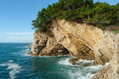 Katmanlı kaya oluşumları ile dramatik bir kayalık ve tepesinde büyüyen çam ağaçları, berrak bir gökyüzünün altında mavi denizi gören. 