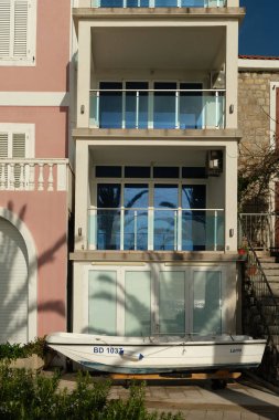 A white boat with blue lettering rests on a wooden stand in front of a modern glass balcony building, reflecting palm trees and sky.  clipart